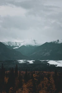 Majestosa cadeia de montanhas com picos cobertos de neve cercada por um vale exuberante e um lago sereno, sob um céu melancólico.