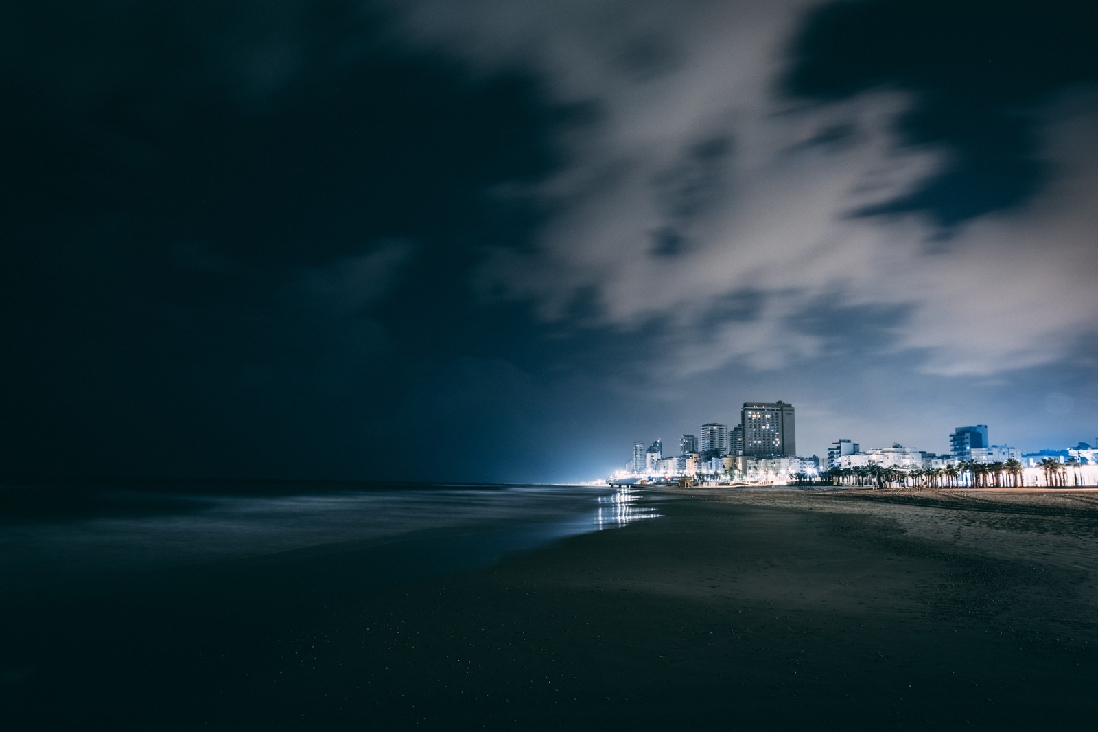 Vista noturna de uma praia com uma cidade ao fundo (noite, azul, nuvem, água, dia)