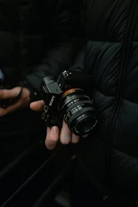 A person holding a Sony digital single-lens reflex camera with a distinctive lens, set against a dark background.