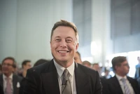 Elon Musk smiling while seated at a formal event, dressed in a suit among an audience.