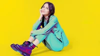 Olivia Rodrigo in a vibrant outfit, seated against a bright yellow background.