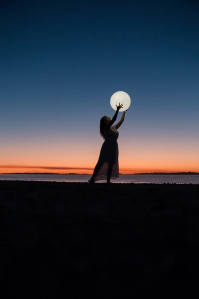 Chica silueteada contra un atardecer, sosteniendo una luna en la playa