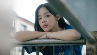 Minji from New Jeans gazes thoughtfully, resting her arms on a railing in an urban setting.