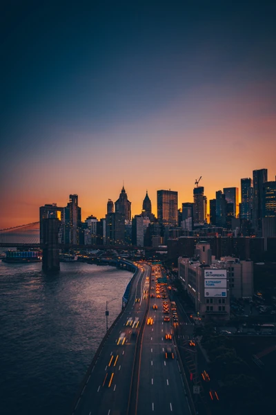 Dusk cityscape featuring the Brooklyn and Manhattan Bridges with skyscrapers silhouetted against an orange and purple horizon.