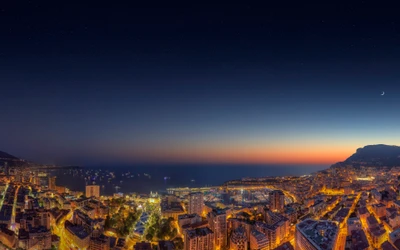 Stadtansicht von Monaco bei Dämmerung: beleuchtete Straßen und ein Halbmond über dem Ozean.