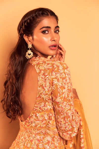 Portrait élégant de Pooja Hegde dans une magnifique tenue brodée, mettant en valeur sa beauté sur un fond chaud.