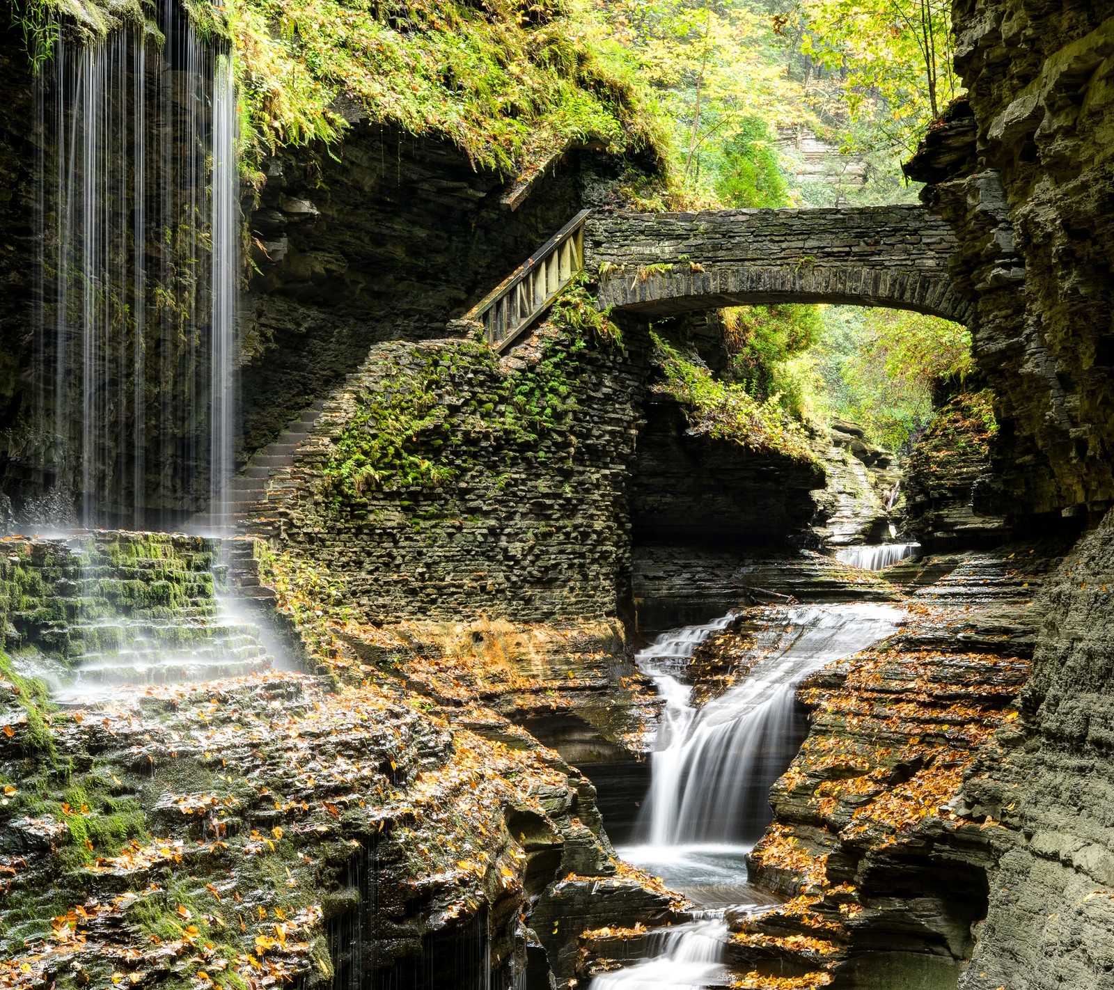 Una cascada fluyendo debajo de un puente en un desfiladero rocoso con un puente sobre él (puente, montaña, roca, agua, cascada)