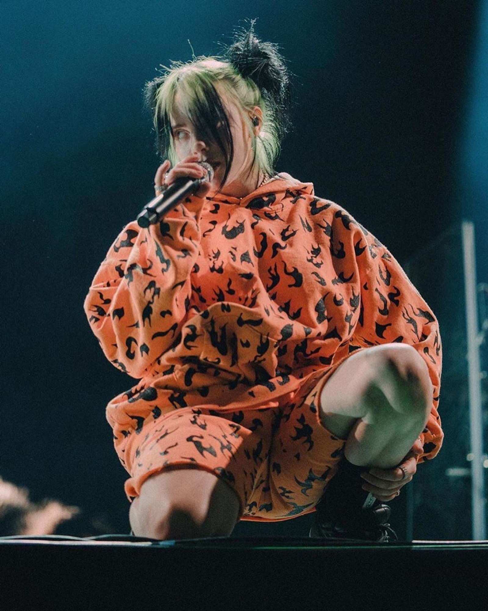 Eine frau in orange outfit, die mit einem mikrofon auf der bühne sitzt (billie, billie eilish, billieeilish, eilish)