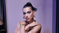 Dua Lipa in a striking pose, showcasing her bold makeup and elegant accessories against a soft purple backdrop.