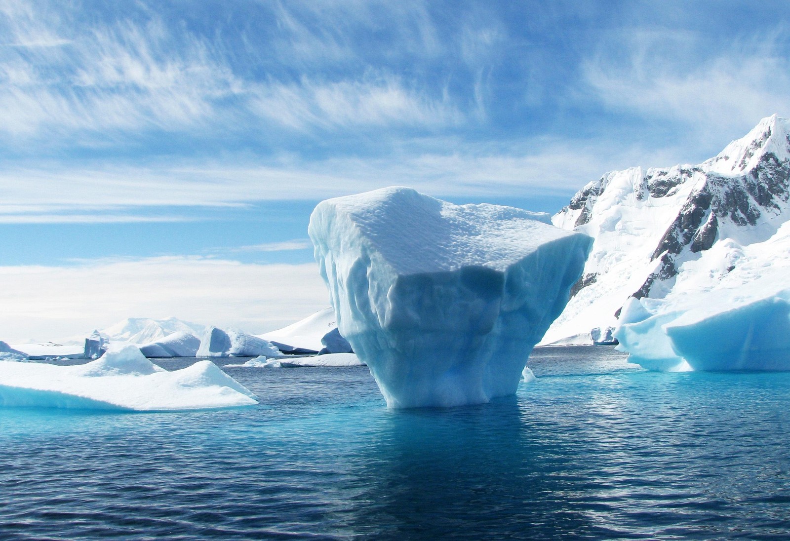 There is a large iceberg floating in the water with a mountain in the background (iceberg, arctic, sea ice, ice, polar ice cap)