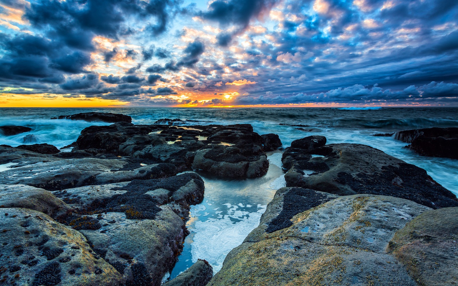 Um pôr do sol sobre o oceano com pedras e água (nuvens tempestuosas, costa rochosa, paisagem marinha, ondas, por do sol)