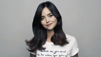 Jenna Coleman in einem weißen T-Shirt mit einer motivierenden Botschaft, die ihr charakteristisches Lächeln und ihre langen, dunklen Haare zeigt.