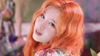 Sana of TWICE with vibrant orange hair, captivatingly gazing back with a playful expression, featured in a colorful setting from the "More & More" album.