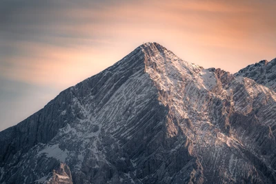 Majestätischer Berggipfel, beleuchtet vom sanften Licht der Morgendämmerung, vor einem ruhigen, wolkengefüllten Himmel.