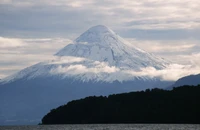 Snow-capped stratovolcano rising majestically above a lush landscape, framed by a dramatic sky and enveloped in soft clouds.