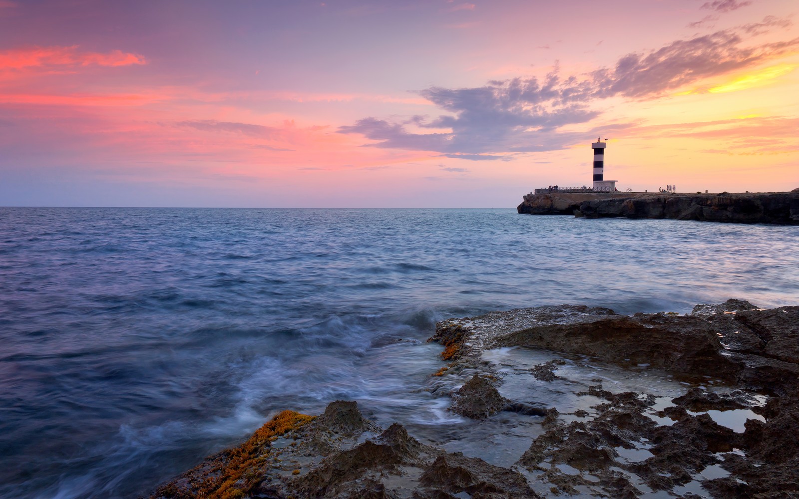 Pôr do sol sobre o oceano com um farol ao longe. (colônia sant jodi, por do sol, costa rochosa, farol, ilha de maiorca)