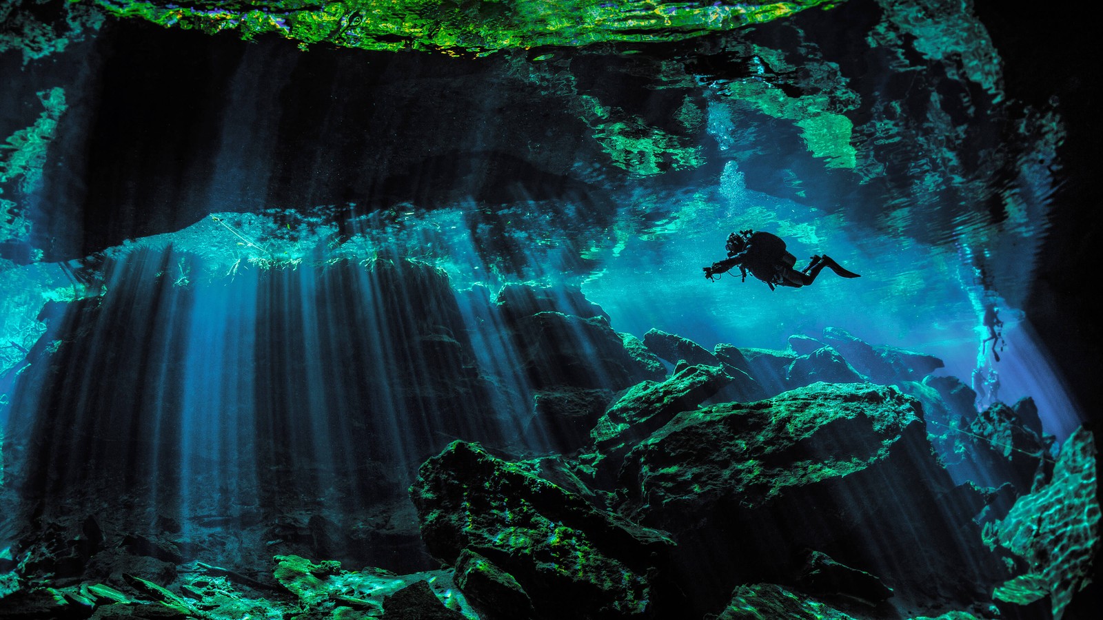 A person swimming in a cave with sunbeams coming out of it (scuba diver, underwater, under the sea, scuba diving, sun rays)