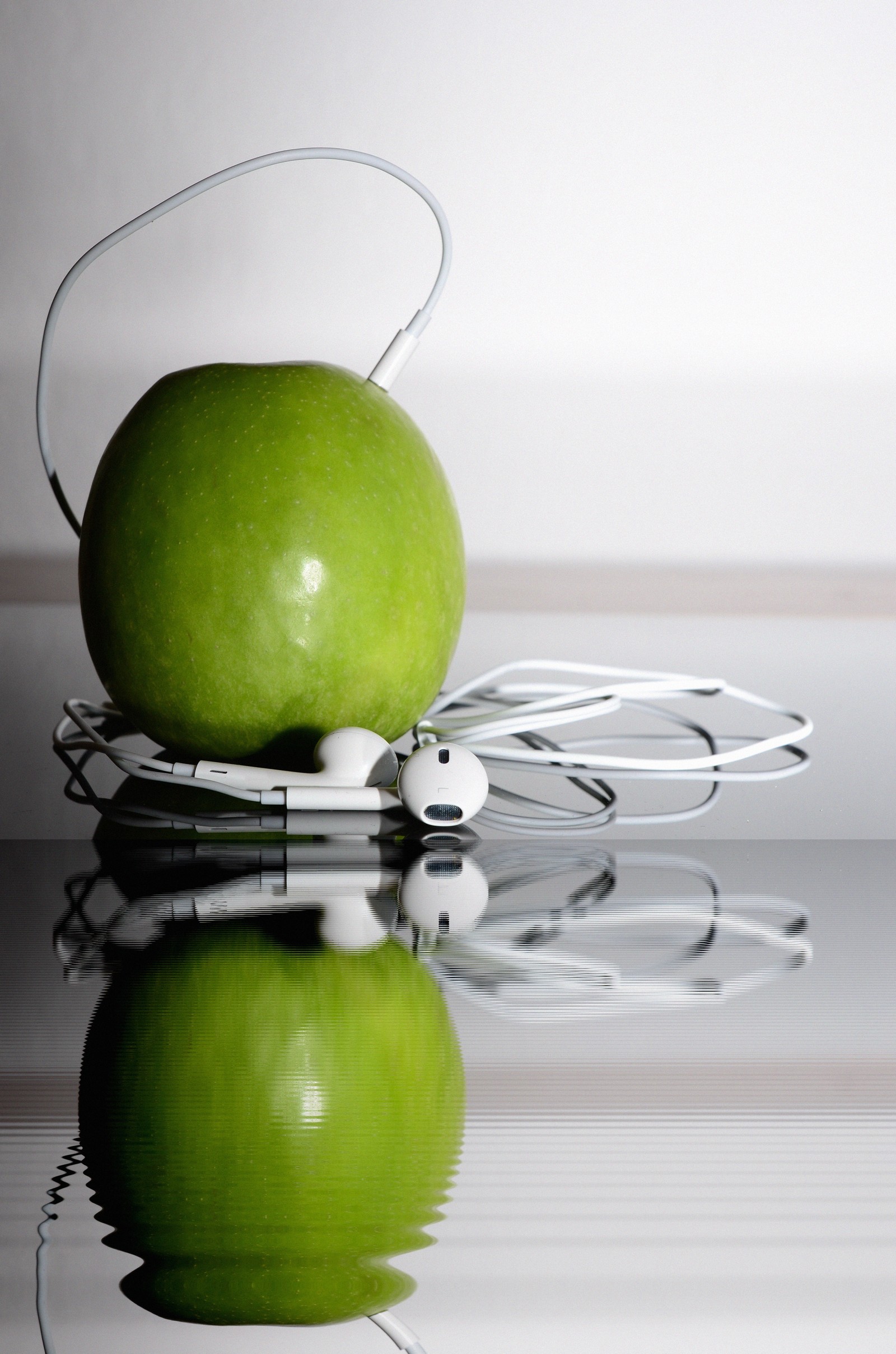 There is a green apple with headphones on a reflective surface (apple earbuds, headphones, green, granny smith, apple)