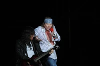 Axl Rose actuando apasionadamente en un concierto de Guns N' Roses, con un guitarrista acompañándolo bajo el cielo nocturno.