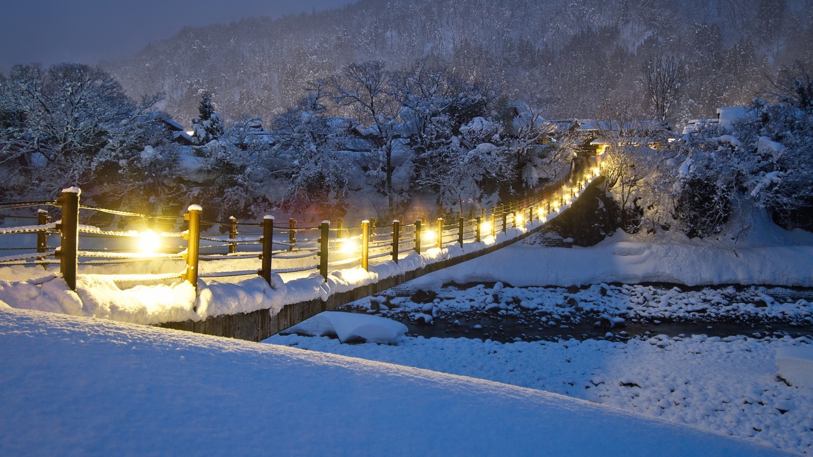 Cena noturna nevada de uma ponte sobre um riacho em um parque (neve, inverno, congelamento, luz, geada)