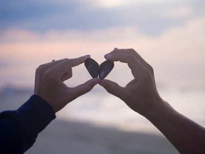 Two hands forming a heart shape with seashells against a serene sky, symbolizing love and connection.