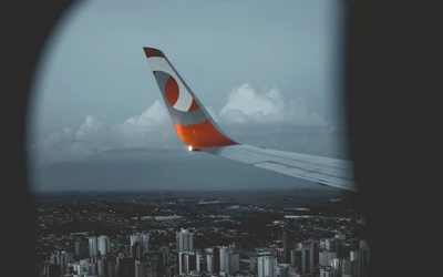 View of an airplane wing with a vibrant tail design above a cityscape, capturing the essence of air travel amidst a cloudy sky.