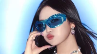 Ningning d'Aespa présentant une déclaration de mode audacieuse avec des lunettes de soleil bleues frappantes et des accessoires glamoureux sur un fond vibrant.