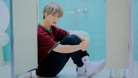 Choi Beomgyu von TXT in einem nachdenklichen Moment, zwischen Toilettenkabinen in einer lebhaften, pastellfarbenen Umgebung sitzend.