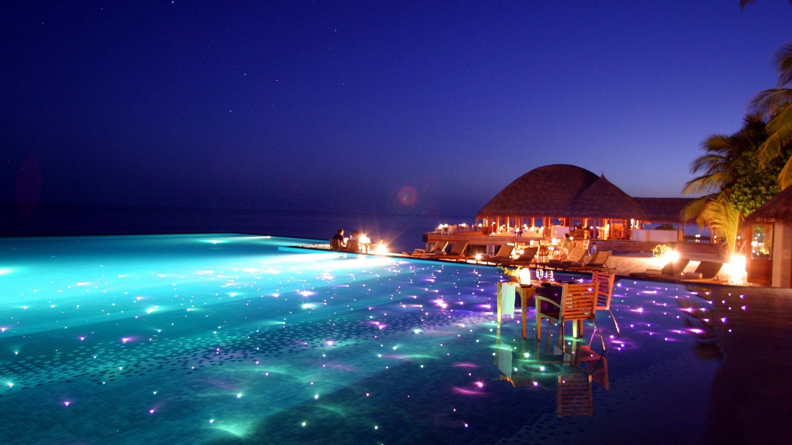 Vista noturna de uma piscina com um bar e espreguiçadeiras ao lado (piscina, estância, noite, iluminação, mar)