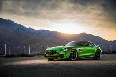 Atemberaubender grüner Mercedes AMG GT vor einer dramatischen Bergkulisse mit Windkraftanlagen bei Sonnenuntergang.