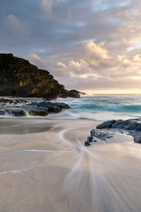 Pôr do sol sobre uma praia tranquila com ondas suaves batendo contra um promontório rochoso, emoldurado por um céu dramático.