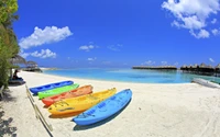 Caiaques coloridos alinhados em uma praia de areia limpa sob um céu azul claro, com águas turquesa tranquilas e bangalôs sobre a água ao fundo.