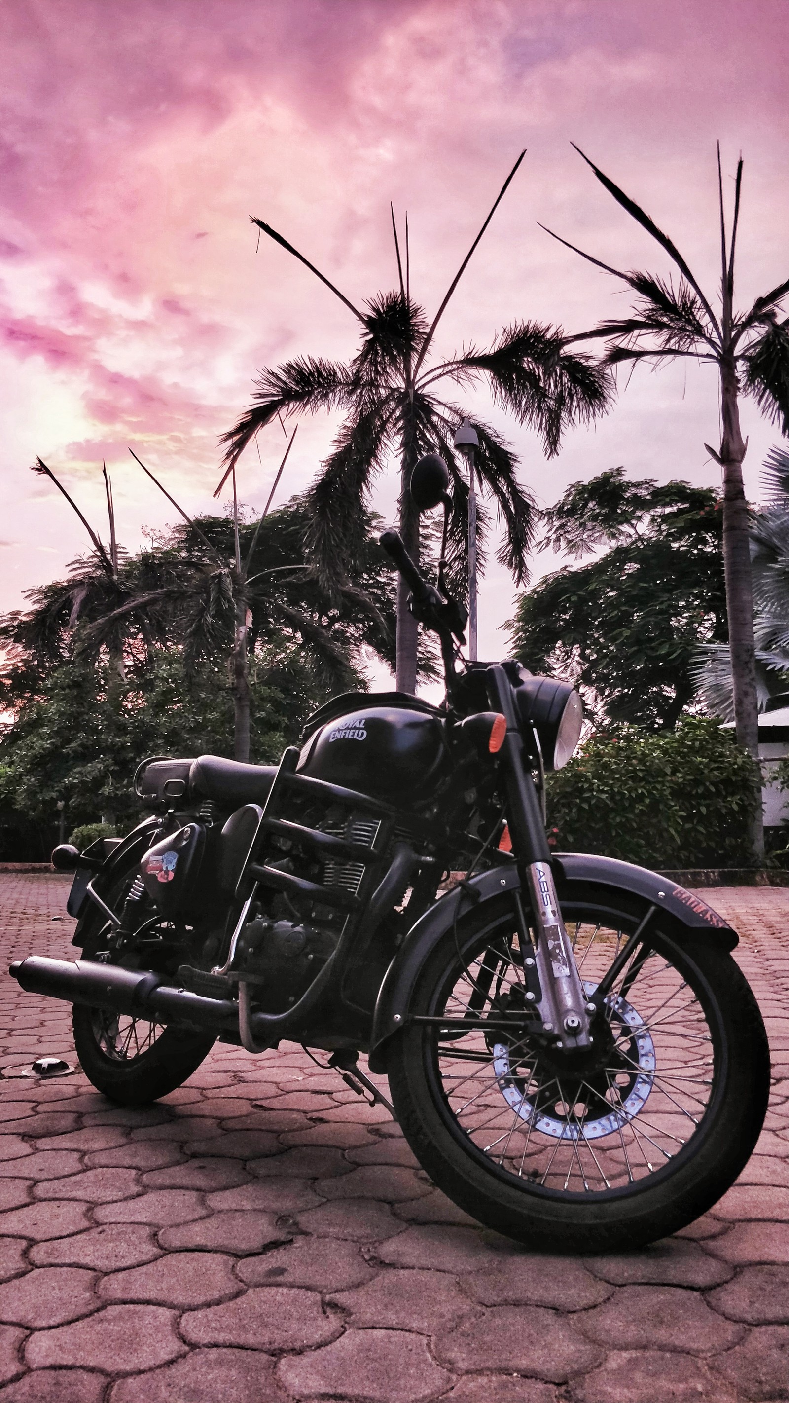 There is a motorcycle parked on a cobblestone road with palm trees (bike, black, classic 500, enfield, royal enfield)