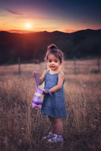 Adorable girl in a field at sunset, holding a colorful bag.