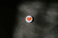 A close-up of a round button featuring an orange heart symbol on a textured dark background.