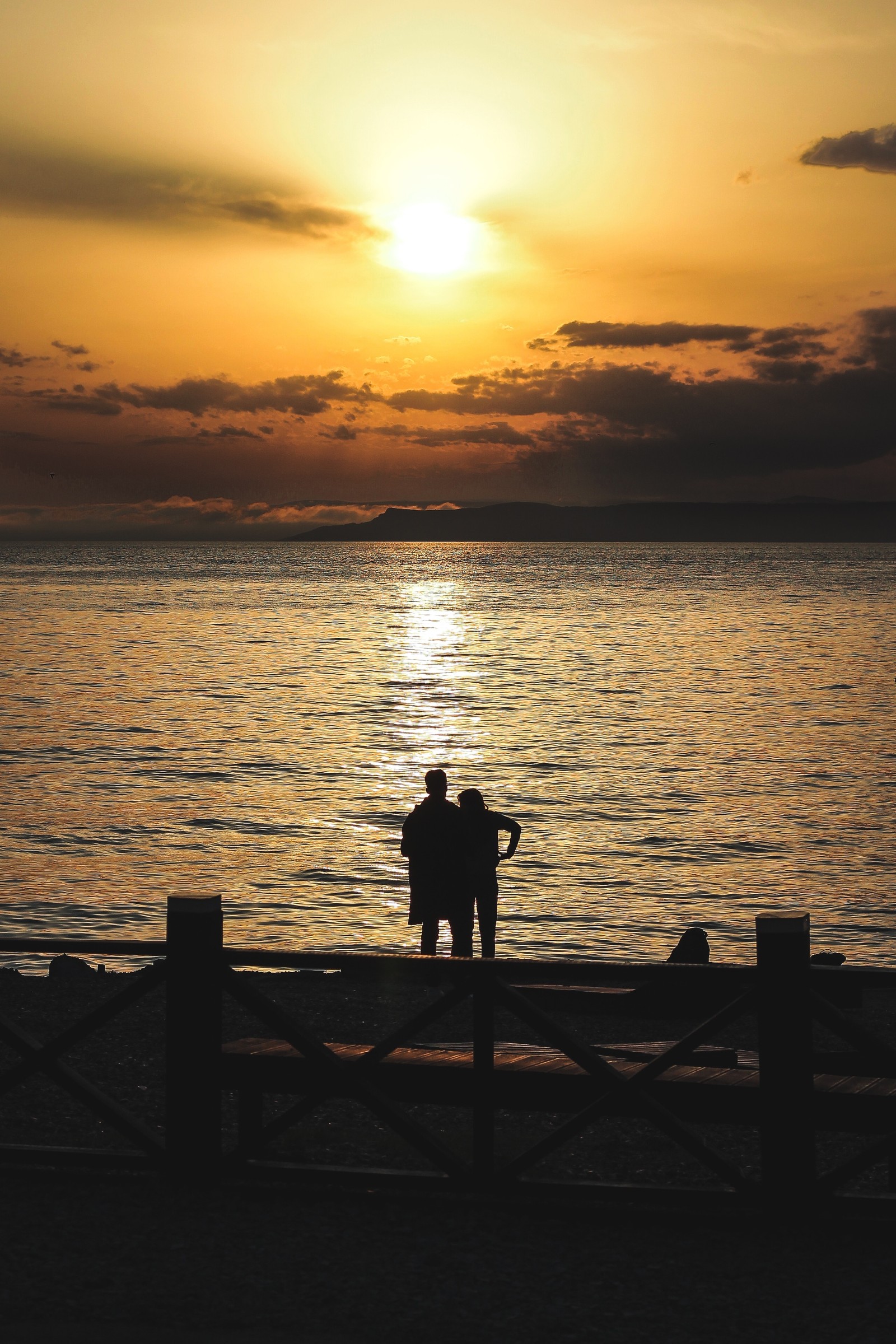 There is a man and a woman sitting on a bench watching the sunset (sunset, silhouette, horizon, sea, ocean)