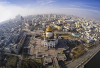 Aerial view of a grand cathedral with golden domes, surrounded by a sprawling urban landscape featuring historic buildings and modern skyscrapers.