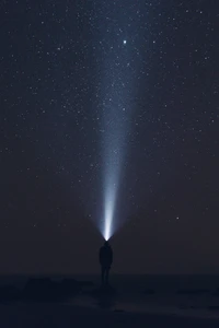 Una figura solitaria en la oscura orilla, iluminada por un rayo de luz que atraviesa el estrellado cielo nocturno, capturando la tranquilidad del cosmos.