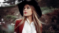 Portrait élégant d'un mannequin de mode avec de longs cheveux bruns flottants, portant un chapeau élégant et un blazer rouge, entouré d'un décor naturel.