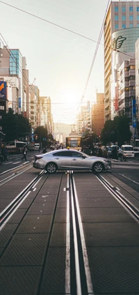 Silver car on tram tracks in a bustling city street at sunset.