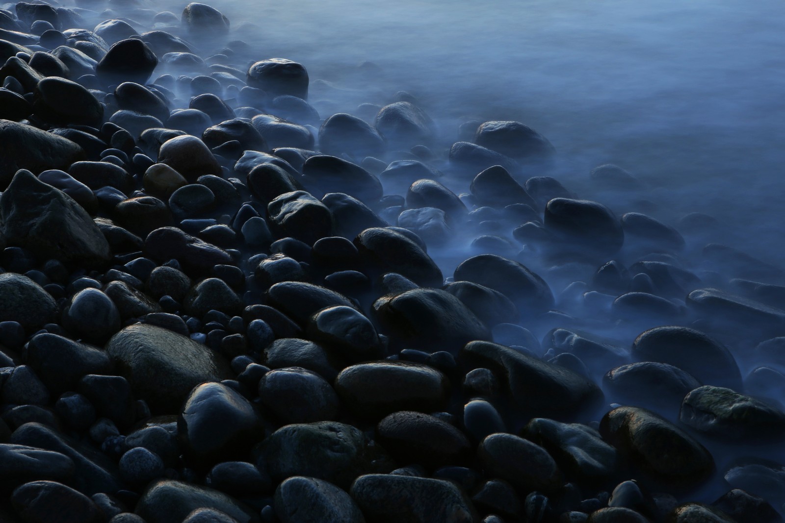 Rocks on the shore of a body of water with a boat in the distance (pebbles, foggy, stones, seashore, mist)