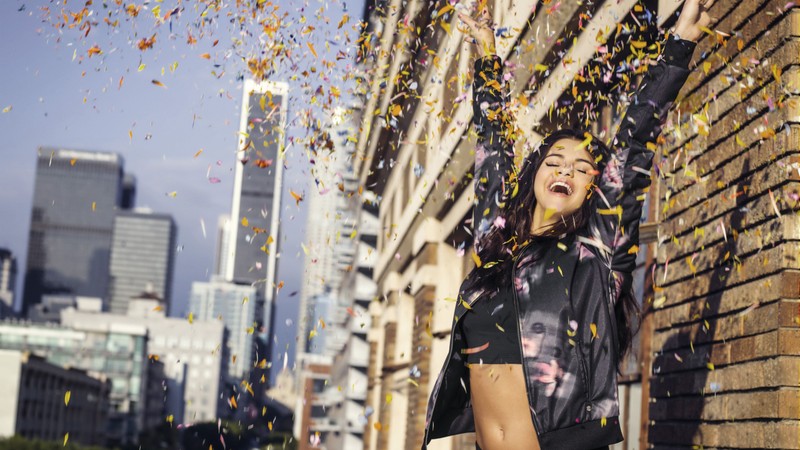 Mujer con chaqueta negra lanzando confeti al aire (selena gomez, edificio, iluminación, feliz, diversión)