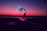 Un homme en silhouette se tient sur le rivage au coucher du soleil, tenant une bombe fumigène contre un ciel rose vibrant, créant une scène esthétique du soir.