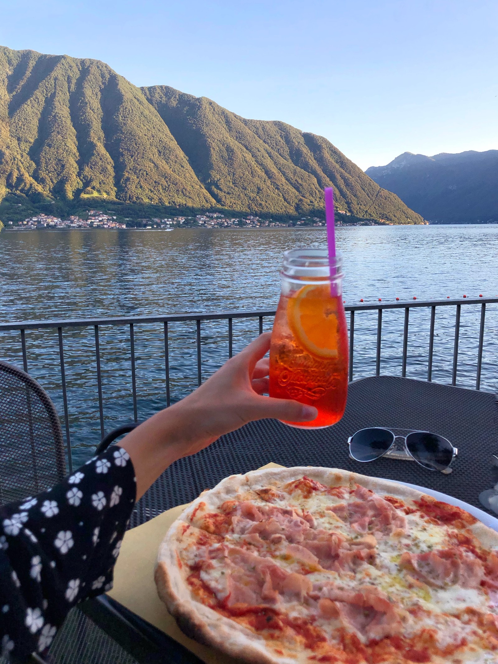 Jemand hält ein getränk und eine pizza auf einem tisch am see (pizza, wasser, gewässer, gebirgige landformen, essen)