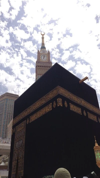 The Kaaba at the Grand Mosque in Mecca, with the clock tower towering in the background under a cloudy sky.