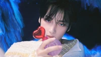 Huening Kai from TXT holds a heart-shaped object, exuding a dreamy and enchanting atmosphere amidst a vibrant, surreal backdrop.