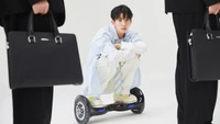 Heeseung from ENHYPEN poses on a hoverboard, showcasing a mix of playful and sophisticated styles while surrounded by figures in formal attire.