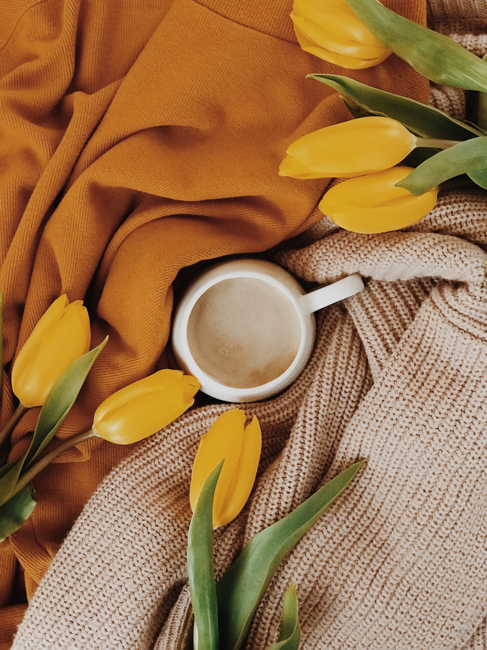 There is a cup of coffee and some yellow tulips on a blanket (yellow, flower, plant, spring, still life)