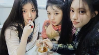 Three young women with distinct hairstyles and fashion styles share a moment at a table, enjoying food together, radiating friendship and youthful energy.