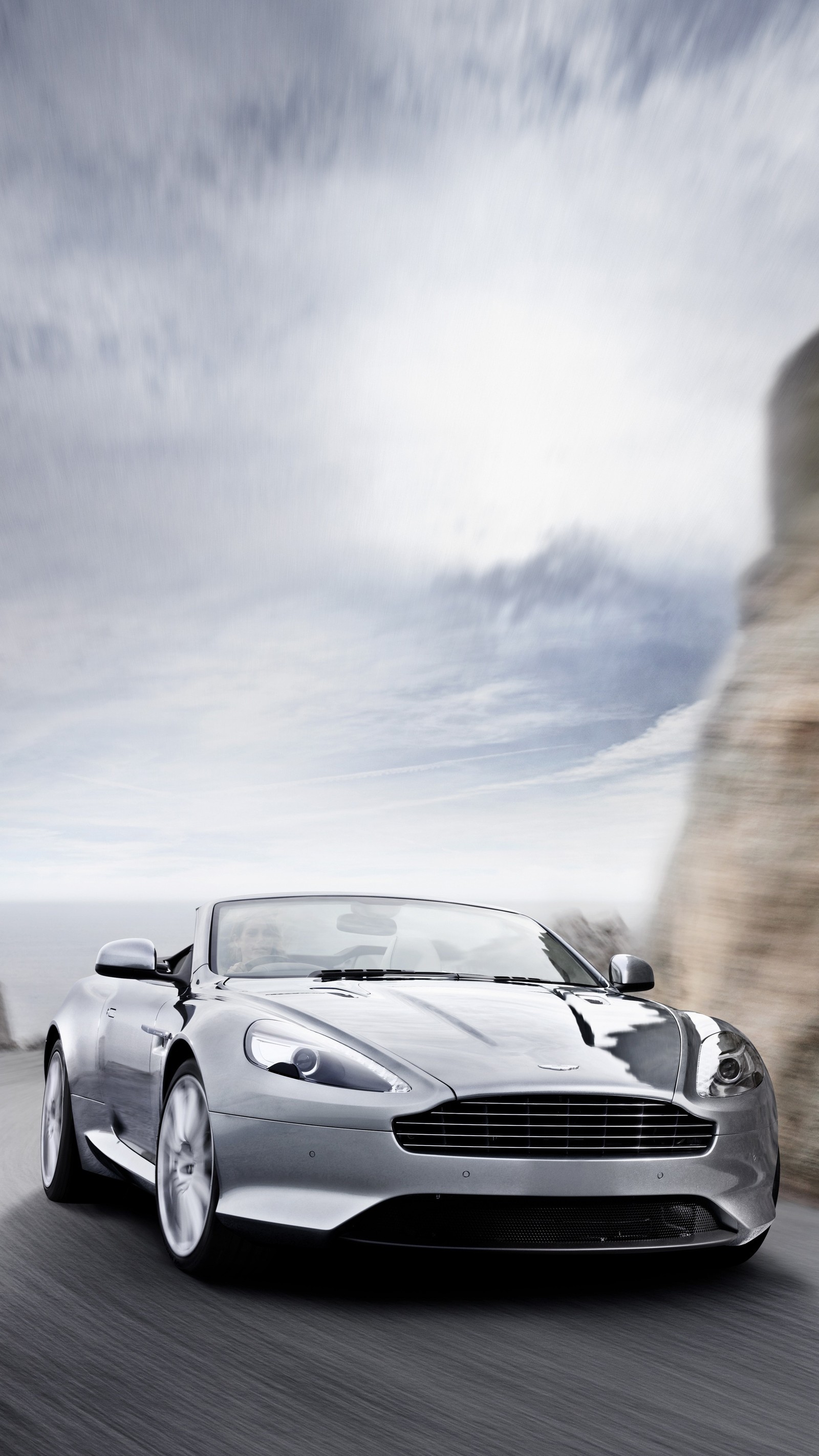 A close up of a silver car driving on a road near a cliff (aston, auto, cars, martin, virage)